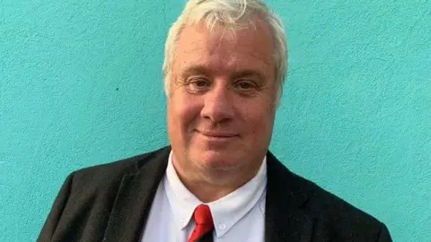 John Wannenburgh, who had short blond hair and is wearing a black jacket over a white shirt and red and black tie.