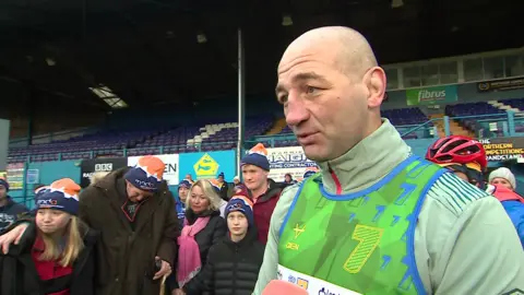 Steve Borthwick at the Fibrus stadium in Workington, being interviewed by a reporter who is out of shot. He is bold and is wearing a green and blue sports vest over a green sports top. Behind him there is a group of supporters wearing MND Association woolly hats.