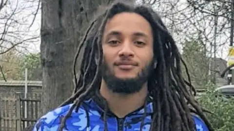 Nimroy Hendricks, who has a short beard and long dreadlocked hair and is wearing a tracksuit top with a blue camouflage design. He is stood in front of a tree, looking gently at the camera.