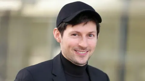 Getty Images Pavel Durov, yang memiliki rambut coklat, mata coklat dan janggut tipis serta mengenakan jas hitam, kaos hitam dan topi baseball hitam, tersenyum ke arah kamera di luar pengadilan Paris untuk sidang