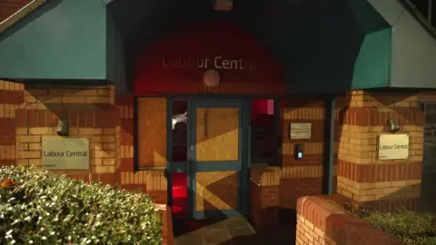 A brick building with a sign over the door reading 'Labour Central. It's glass door is boarded up. It is night time.