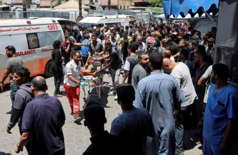 Reuters A person is wheeled towards a hospital, blood visible on their arm, by an ambulance medic, while a crowd gathers round