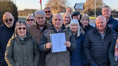 Castle Point Borough Council A group of men and women standing by a road outdoors. At the centre is council leader Dave Blackwell, who is holding a letter in a plastic sleeve.
