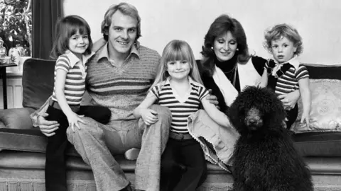Getty Images A black and white image of a family of 3 children and a father and mother. All sat on a couch with a black fluffy dog.
