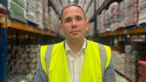 Mr Gamble is standing in a warehouse. He's wearing a yellow high-viz jacket. Behind him are shelves stacked with goods.