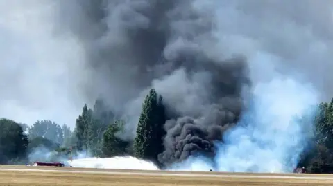 UKNIP A huge billowing cloud of black, grey and white smoke coming from the edge of the airfield. It is so thick that it is hiding some of the trees - that are on the edge of the airfield. A fire engine can be seen spraying water towards the mass of smoke.