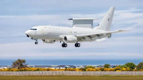 Wedgetail airborne early warning (AEW) plane coming into land, with yellow bushes, houses and the sea in the distance.