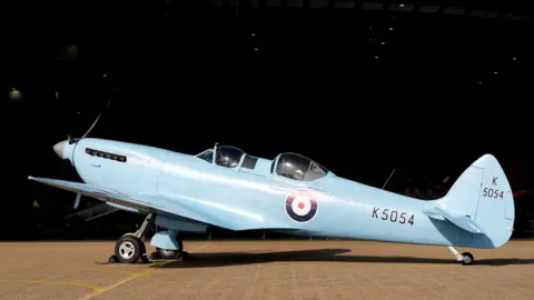 A two-seater Spitfire painted light blue with the RAF logo and "K5054" written in black towards the back of the aircraft. The sky is black.