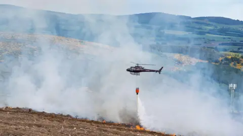 Jasperimage A helicopter waterbombs flames in a wildfire on a barren hillside. Water pours from a large red bucket under the helicopter.
