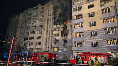 Reuters Sees smoke from a burning residential building in Hlevkha, Kiev region, Ukraine, after a Russian drone strike. Emergency workers stand outside a fire engine.