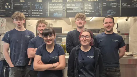Six members of staff stand in a fish and chip shop with dark T-shirts which say "The Fisheries". The store counter and menu can be seen behind them. 