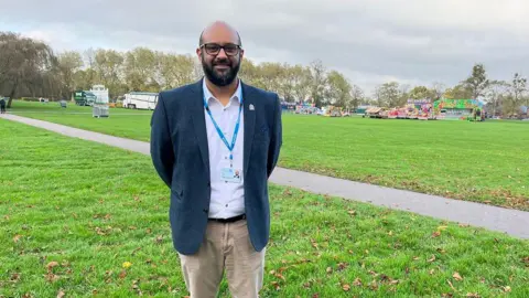 Local councilor Bal Anota stands in Walks Community Park in King's Lynn. He is wearing a blue jacket, white top and light brown chinos. 