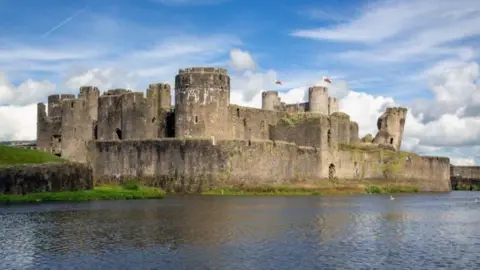 Getty Images Caerphilly Castle with a blue sky and clouds overhead, overlooking a moat. Two Welsh flags can be seen flying from the building.