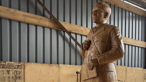 The sculpture of JRR Tolkien carved from oak stands in a warehouse. He is depicted in his army uniform and is holding a notebook