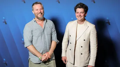 Getty Images Two men stood in front of a blue background, smiling and looking at the camera. The man on the left has grey hair and a beard and is wearing a light grey linen shirt and a beaded brown necklace. The man on the right has curly dark hair and is wearing a cream t shirt and suit jacket.