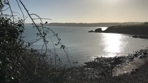 A cove is seen with the sun shimmering on the sea and a rock formation in the background 