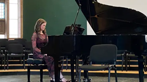 Susan Terris Freya Terris sits at a grand piano, wearing a long sleeved dark dress with red flowers, and black strappy heels. The room behind her has green walls and large, long windows. She is also surrounded by chairs.