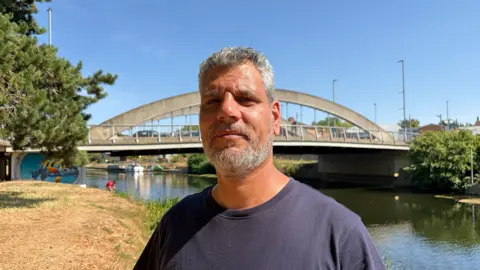 Samer Jouha standing by the River Avon in Evesham, close to where he pulled the woman from the water. It is a sunny day and behind him is a bridge. There are also trees behind him on both banks of the river.
