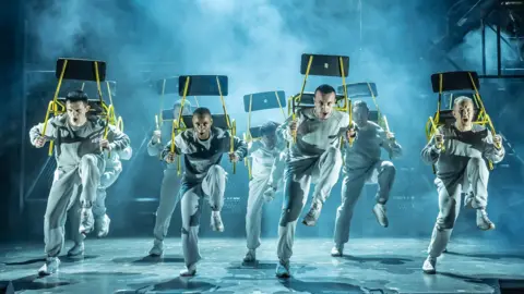 Marc Bennett A group of performers in white outfits dance in formation during a production of an Officer and a Gentleman at the Bristol Hippodrome. They are looking stern and carrying chairs on their backs