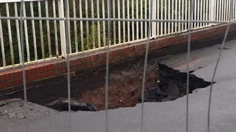 A hole in the road surface, with white railings and brickwork above the hole. The asphalt has been cracked open, and soil is visible. There are trees in the distance and security fencing in the immediate foreground through which the photo was taken.