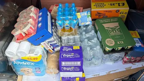 Pritchard Brothers A number of boxes containing foods like porridge, chocolate fingers, Pot Noodles, and Doritos, as well as bottles of water and Lucozade. There is also a multipack of toilet roll.