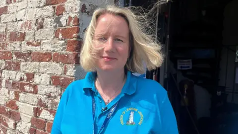 A woman with blonde hair and wearing a light blue t-shirt is standing at the entrance to the lighthouse.