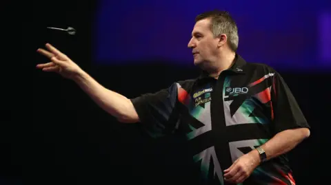 Getty Images Ronnie Baxter seen side on throwing a dart with his right hand. He is wearing a black shirt with a black and white Union Flag on it.