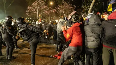 EPA-EFE/REX/Shutterstock Riot police clash with protesters at night. One officer in riot gear kicks a protestor who has his hands to his face.