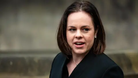 Getty Images Head-and-shoulders shot of Kate Forbes, who has shoulder-length brown hair, looking off to her left. She is wearing a black blazer-style jacket and standing outside a grey church building. Her mouth is open as if she is talking to someone.