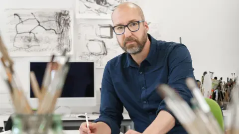 University of Sunderland Nick Lewis looks at the camera while posing with a pencil in his hand. He is wearing a navy shirt and glasses, is bald with a dark and grey beard. Behind him is a computer and several charcoal sketches of shapes on the wall.