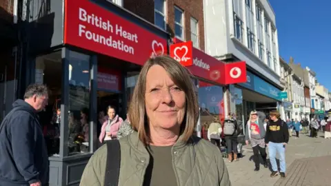 Andrew Turner/BBC Jane Hare is pictured standing outside the British Heart Foundation shop, which had a red sign on the front with white lettering. She wears an olive green quilted jacket and a matching knit top. She has a black bag strap over her right shoulder and has shoulder-length light brown hair. Other people are walking near her.