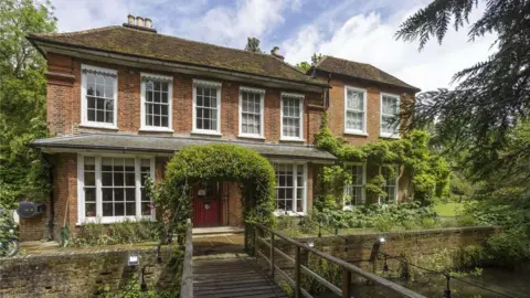 Savills The front of a house with several windows in front of a small pedestrian bridge over a mill stream