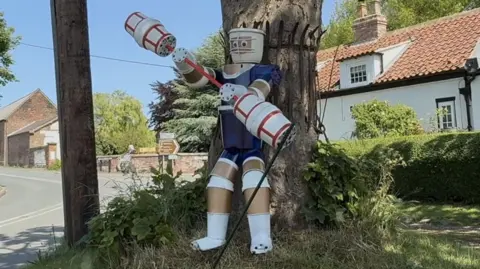 BBC/Amber Gash A character from Gladiators stands on a grass mound under a tree. The statue is holding a large stick used in one of the contests.