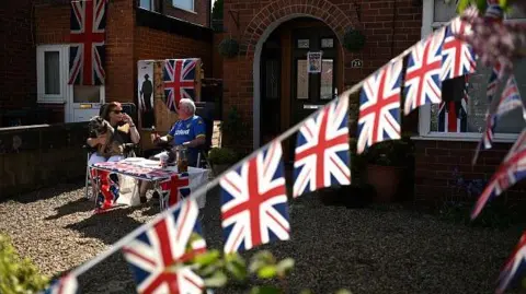 Getty Images Two people have a VE Day tea party in a front garden decorated with Union flags during the Covid pandemic, when social distancing was in place. It is sunny and they are sitting at a table. The woman is wearing sunglasses.