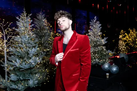 Tom Grennan mengenakan jaket satin merah dan berpose di samping deretan Pohon Natal saat syuting Top Of The Pops edisi Natal 2024