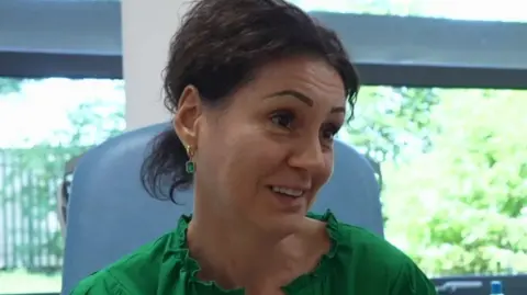 A close-up of Madalina Chifu. She is sitting in a chair in a clinical setting, and is wearing a green top and has short dark hair partially tied back.