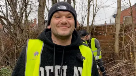 Dan Bater/BBC Man with a black hat, a wollen hoodie and a bright yellow hi-vis. Background of bare tree, bushes and a litter picker in hi-vis behind him.