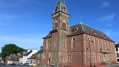 Leslie Barrie The Wigtown County Buildings a prominent brick building with a tower and a clock on it at the end of a row of houses with a blue sky overhead
