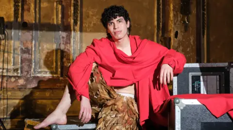 Jacob Alon, wearing gold feathered trousers and an oversized red sweater, poses on flight cases backstage at the BBC's Jools Holland show