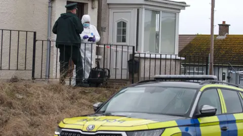 A police man and person in white PPE gear are standing outside a white pebble dashed house. A police car is parked in the foreground