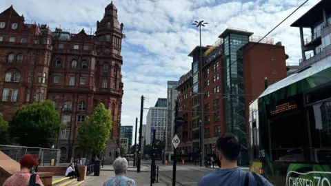 Scene of Manchester city centre. Two women and a man walk along the street as a bus passes them by.
