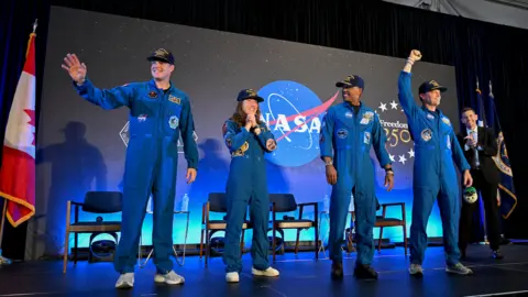 This indoor photograph shows the four Artemis II crew members standing on a stage in matching royal-blue flight suits. Behind them is a large dark backdrop with a prominent NASA logo and starry design, with national flags placed at the sides. One astronaut waves, another appears to speak into a microphone, and another raises a fist in celebration. The lighting is bright and theatrical, and the mood is triumphant, as if the crew are being welcomed back after their mission.