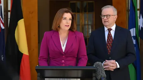 EPA A woman in a magenta jackets stands in front of a podium with a man in a suit and tie to her left.