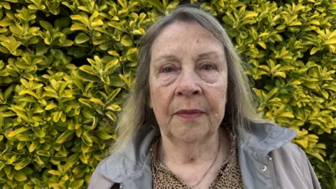 Woman in grey jacket stands in front of yellow/green hedge