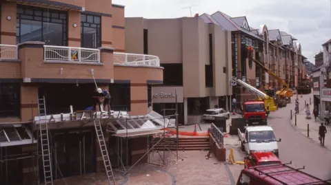 Barnsley Archives A street scene where construction or renovation work is taking place on a row of commercial buildings. In the foreground, there is a building with a curved facade made of light brown brick and large glass windows. Two workers are standing on scaffolding and a ladder, installing or repairing a metallic sign that reads “ALHAMBRA” above the entrance. The area around the entrance is partially cordoned off, and construction materials are visible.
Further down the street, additional buildings with contemporary architectural designs can be seen, featuring beige and brick exteriors with sloped roofs and dormer windows. Several vehicles, including red and white trucks and a yellow crane, are parked along the street, and another crane is lifting a worker to an upper level of one of the buildings. Pedestrians are walking on the sidewalk in the background. The sky is overcast.