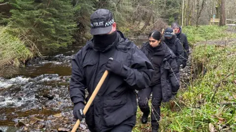 Empat petugas polisi berpakaian hitam berjalan di sepanjang sungai. Mereka memegang tongkat kayu dan melihat ke bawah.