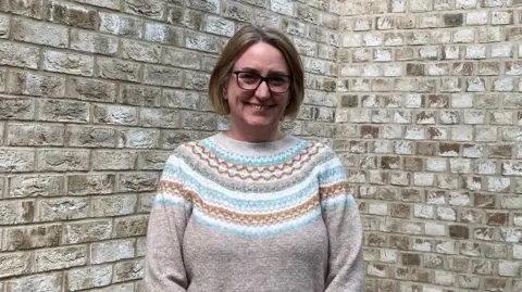 Emma Howgego/BBC Vicky Jenner stares at the camera. She wears black rimmed glasses, a beige jumper with a blue, white and gold pattern on the top half. She stands in front of a brick wall.