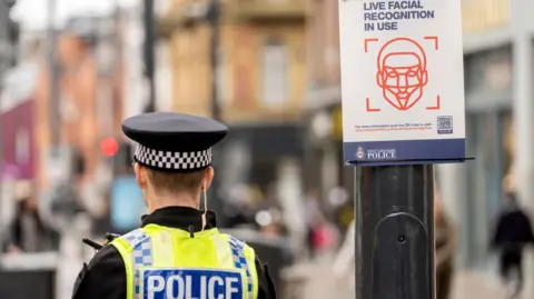 PA Media A police officer stands with his back next to a sign fixed to a lamp post in a busy street. The sign has a graphic of a face and says: Live facial recognition in use.