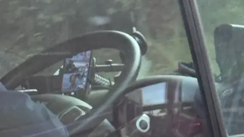 a driver at a black steering wheel with a phone mounted on the dashboard with tiktok on the screen.