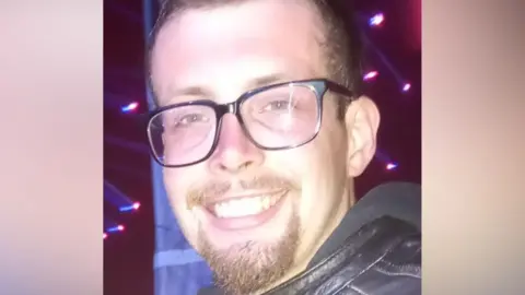 A man with short brown hair and a short brown goatee. He is wearing black framed glasses and a black leather jacket.